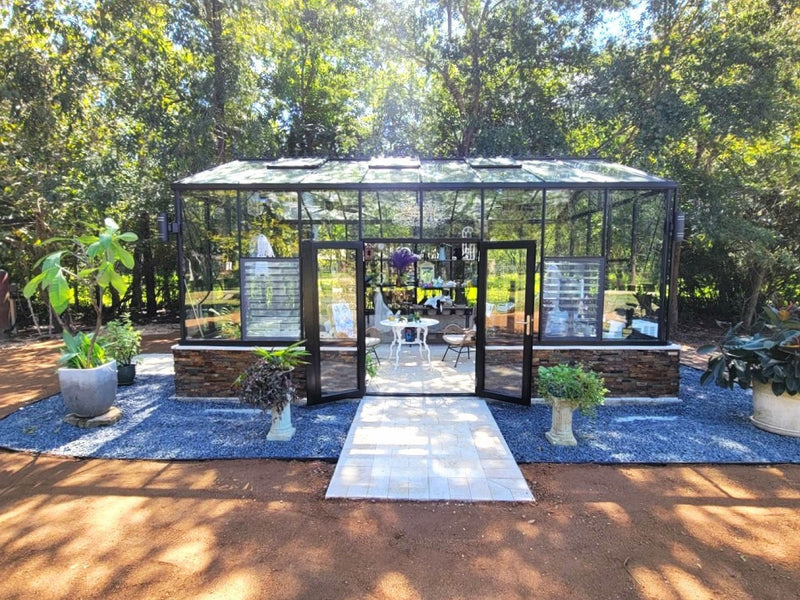 Royal Victorian VI36 greenhouse with double doors open in a wooded garden, set on gravel with a paver walkway