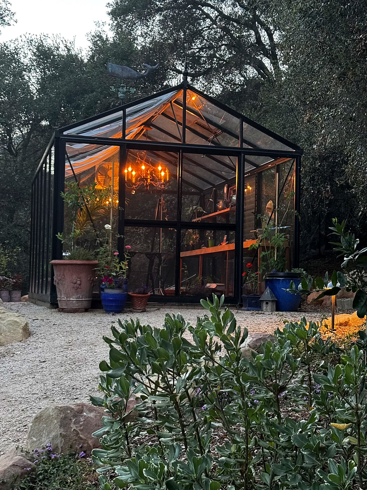Customer photo: black Royal Victorian VI34 glass greenhouse at dusk, lit by a chandelier inside, on a gravel garden path