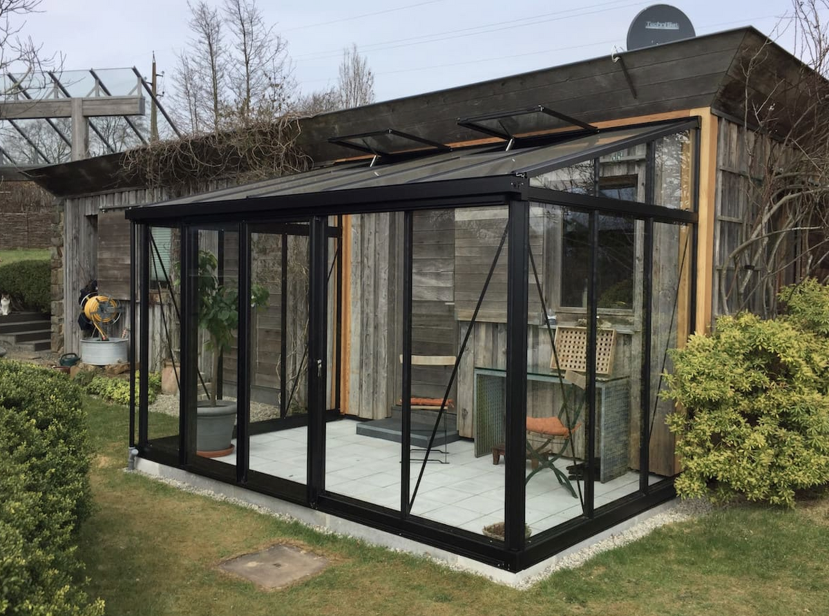 Black Janssens Arcadia lean-to greenhouse with sliding doors beside a garden building