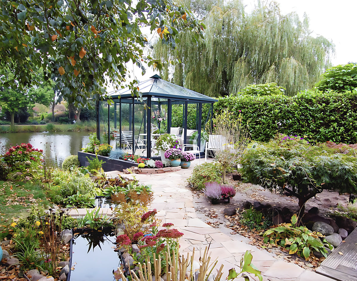Janssens Pyramid glass greenhouse with black frame beside a pond in a landscaped garden