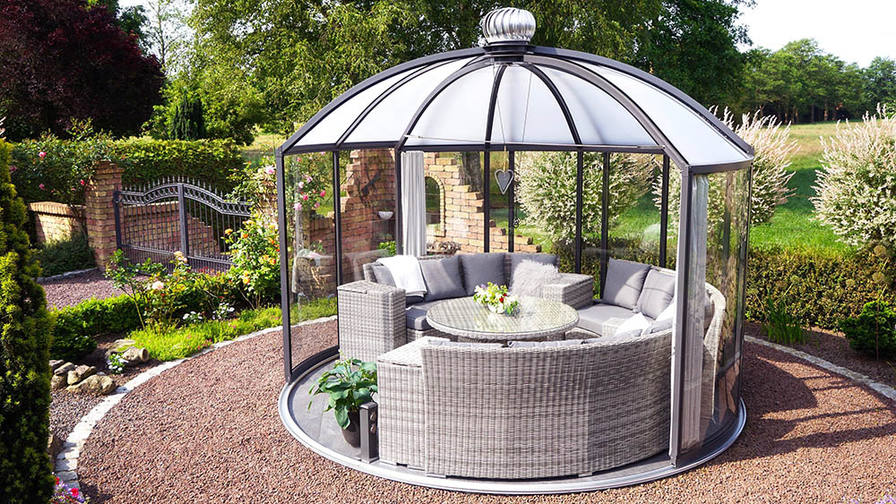 Hoklartherm Rondo glass garden pavilion with dome roof and dark grey frame on a gravel circle, furnished with a wicker lounge set