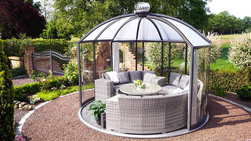 Hoklartherm Rondo glass garden pavilion with dome roof and dark grey frame on a gravel circle, furnished with a wicker lounge set