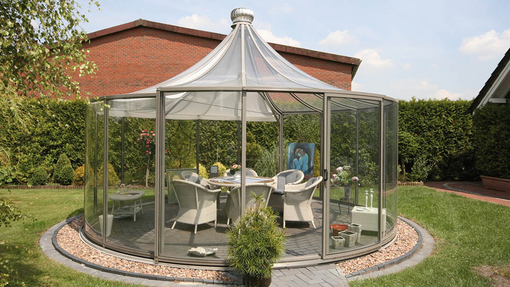 Rondo round glass pavilion with pyramid roof and silver-grey frame on a lawn beside a brick house, set for dining