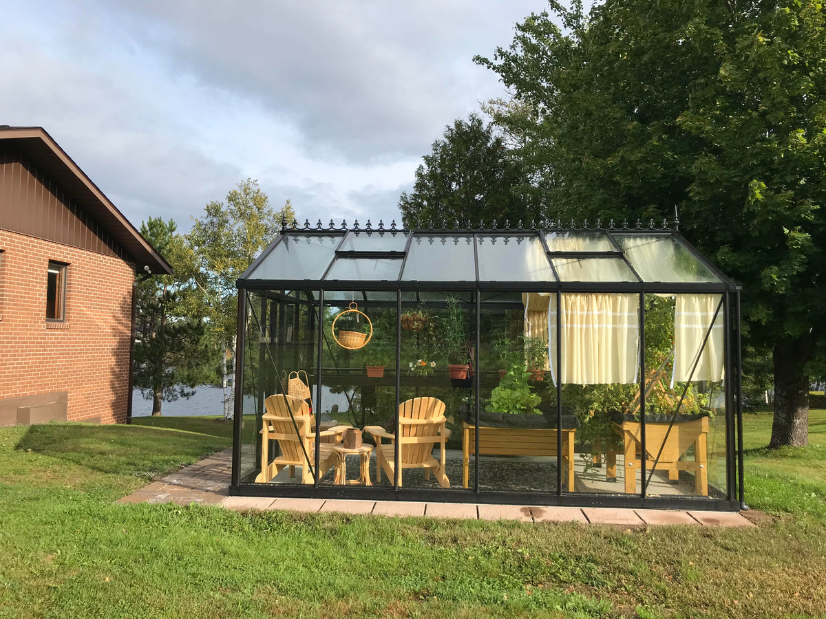 Royal Victorian VI34 greenhouse on a paver base on a lawn, furnished with wooden chairs and plants